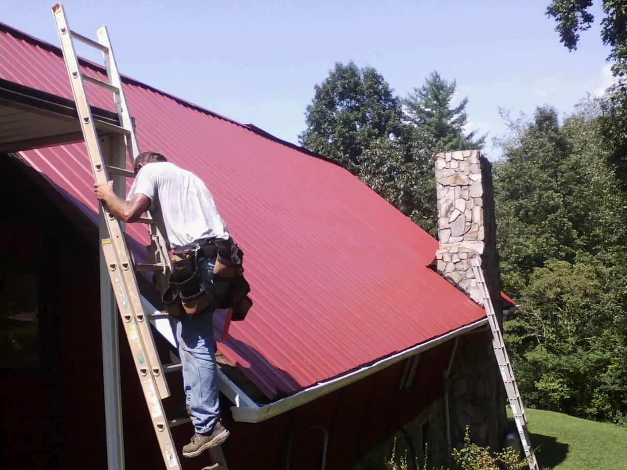 Roofer on a ladder beside a red metal roof and stone chimney — Soffit & Fascia Repair by Roofing Brothers in New Cambria